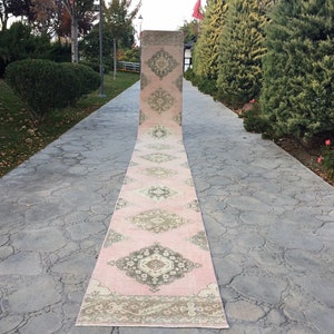 May include: A long, pink and cream runner rug with a traditional Persian design. The rug is laid out on a gray concrete walkway.