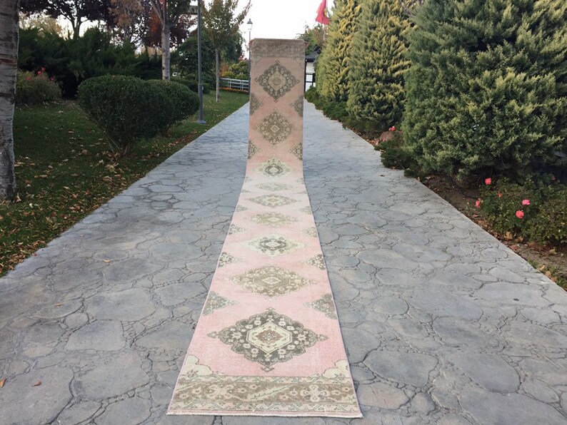 May include: A long, pink and cream runner rug with a traditional Persian design. The rug is laid out on a gray concrete walkway.