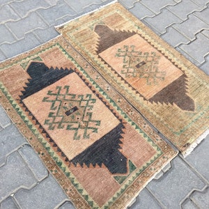 May include: Two small, rectangular, hand-woven rugs with a geometric pattern in shades of brown, green, and beige. The rugs have a fringe border.
