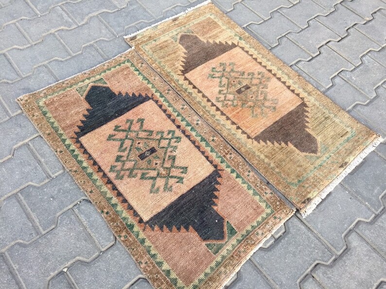 May include: Two small, rectangular, hand-woven rugs with a geometric pattern in shades of brown, green, and beige. The rugs have a fringe border.
