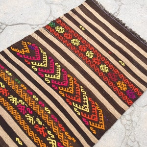 Puede incluir: Una alfombra de rayas marrones y beige con patrones geom&eacute;tricos coloridos tejidos en el dise&ntilde;o. La alfombra tiene flecos en los extremos.