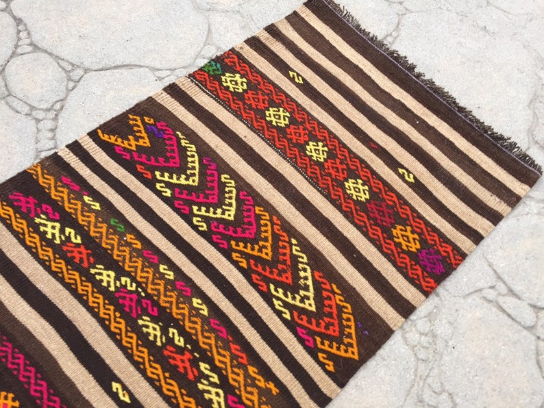 Puede incluir: Una alfombra de rayas marrones y beige con patrones geom&eacute;tricos coloridos tejidos en el dise&ntilde;o. La alfombra tiene flecos en los extremos.