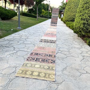 May include: A long, narrow runner rug featuring a series of repeating rectangular panels in muted tones of olive green, dusty pink, cream, and charcoal black. The rug displays intricate geometric patterns and floral motifs across its entire length.