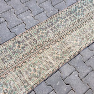 May include: A long, narrow rug with a geometric pattern in shades of green, brown, and beige. The rug is laid out on a gray concrete surface.