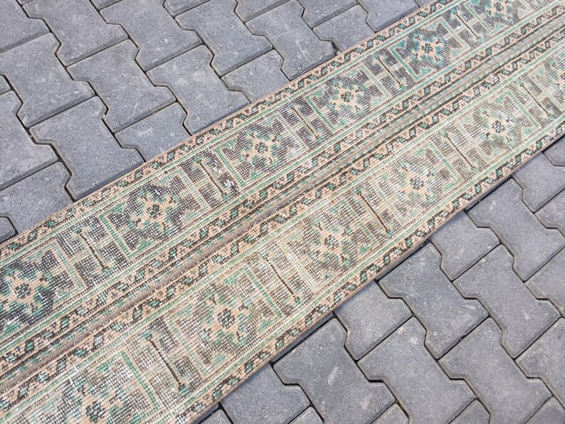 May include: A long, narrow rug with a geometric pattern in shades of green, brown, and beige. The rug is laid out on a gray concrete surface.