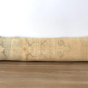 May include: A long, rectangular lumbar pillow featuring a faded, tan-colored woven textile with subtle, muted green and dark gray geometric patterns. The fabric displays a textured, worn appearance with visible weave details across the entire surface.