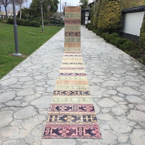 May include: A long, narrow rug with a colorful geometric pattern in shades of pink, beige, and blue. The rug is laid out on a stone path in a garden setting.
