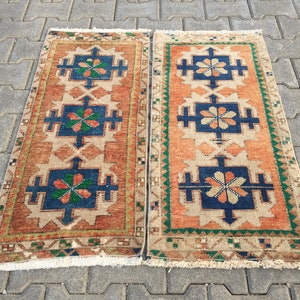 May include: Two vintage Turkish rugs with a geometric pattern in shades of orange, blue, green, and beige. The rugs have a fringe border and are laid out on a gray concrete surface.