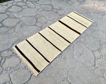May include: A rectangular, cream-colored runner rug with dark brown stripes. The rug is made of woven material and has a fringed edge. The rug is approximately 60 inches long and 24 inches wide.