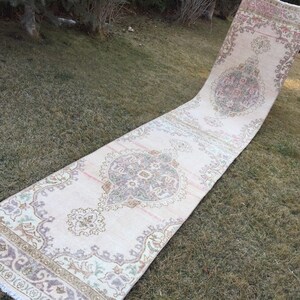 May include: A long, off-white runner rug with a floral pattern. The rug has a fringed edge and is laid out on a grassy surface.