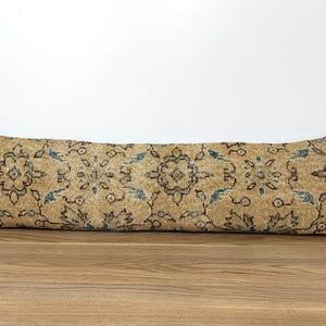 May include: A long, rectangular pillow with a beige background and an intricate floral pattern in shades of brown and blue. The pillow is placed on a wooden surface, showcasing its decorative design.