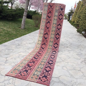 Puede incluir: Una alfombra larga y estrecha, tejida a mano, con un diseño persa tradicional. La alfombra es de tonos rosa, verde y negro. Está doblada por la mitad y puesta sobre un camino de hormigón.