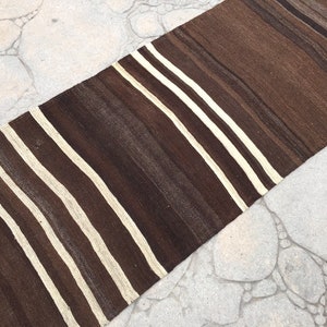 Puede incluir: Una alfombra larga y estrecha de rayas marrones y blancas con textura tejida. La alfombra est&aacute; sobre una superficie de hormig&oacute;n gris claro.