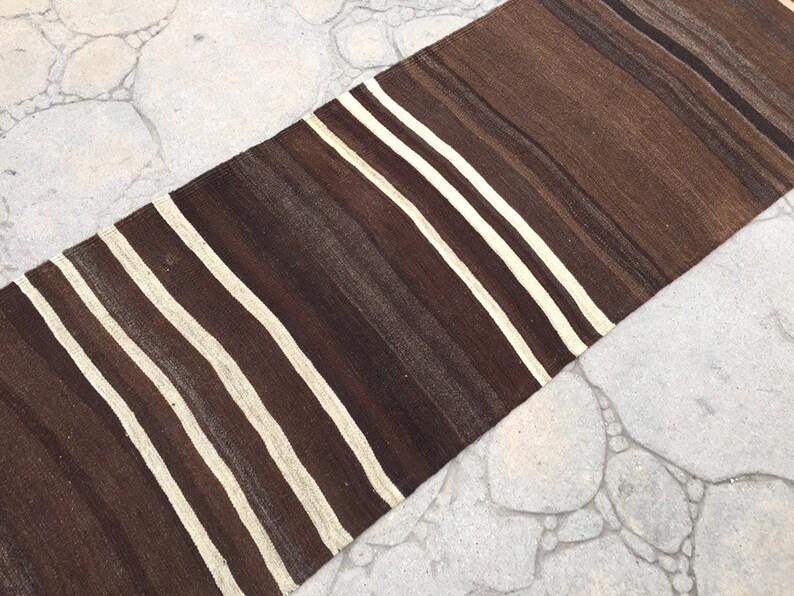 Puede incluir: Una alfombra larga y estrecha de rayas marrones y blancas con textura tejida. La alfombra est&aacute; sobre una superficie de hormig&oacute;n gris claro.