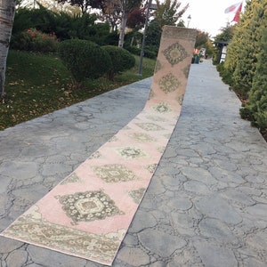 May include: A long, pink and cream runner rug with a geometric pattern. The rug is laid out on a gray concrete walkway.