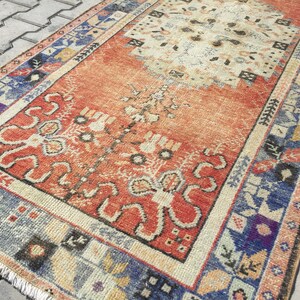 May include: A vintage Turkish runner rug with a red, orange, and blue geometric pattern. The rug has a faded, distressed look and features a central medallion design.