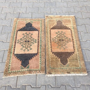 May include: Two vintage Turkish rugs with geometric patterns in shades of brown, green, and beige. The rugs have a fringe border and are laid out on a grey concrete surface.