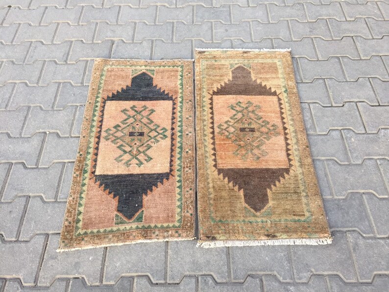 May include: Two vintage Turkish rugs with geometric patterns in shades of brown, green, and beige. The rugs have a fringe border and are laid out on a grey concrete surface.