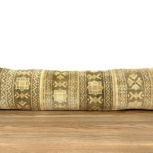 May include: A long, rectangular pillow with a woven design. The pillow features a pattern of repeating geometric shapes in shades of brown, beige, and light blue. The pillow is placed on a wooden surface.