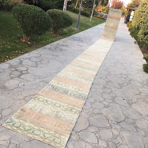 May include: A long, narrow, handwoven rug with a light green, pink, and beige colour scheme. The rug is laid out on a grey concrete walkway.