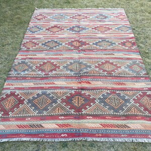 May include: A traditional Turkish kilim rug with a geometric pattern in shades of red, orange, brown, and blue. The rug has a fringed edge and is made of wool.