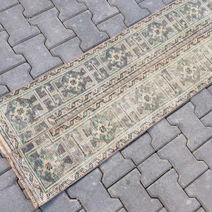 May include: A long, narrow rug with a beige background and a repeating pattern of green and brown geometric shapes. The rug is lying on a gray concrete surface.
