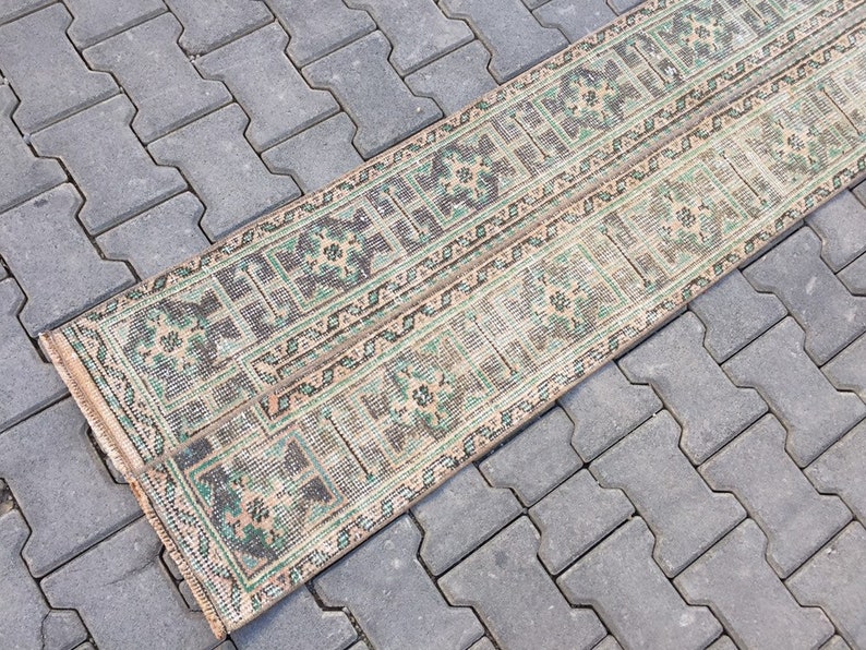 May include: A long, narrow rug with a beige background and a repeating pattern of green and brown geometric shapes. The rug is lying on a gray concrete surface.