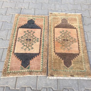 May include: Two vintage Turkish rugs with a geometric pattern in shades of brown, green, and beige. The rugs have a fringe border and are laid out on a grey concrete surface.