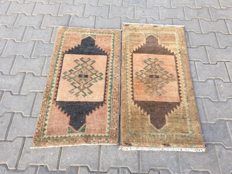 May include: Two vintage Turkish rugs with a geometric pattern in shades of brown, green, and beige. The rugs have a fringe border and are laid out on a grey concrete surface.
