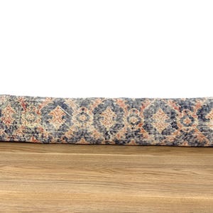 Puede incluir: Un cojín largo y rectangular con un diseño de alfombra vintage. El cojín presenta un patrón de colores azul, naranja y crema. El cojín está colocado sobre una superficie de madera.