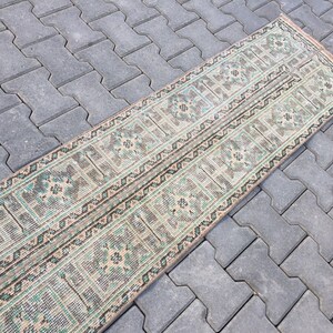 May include: A long, narrow rug with a geometric pattern in shades of beige, green, and brown. The rug has a distressed look, with some areas faded and worn.