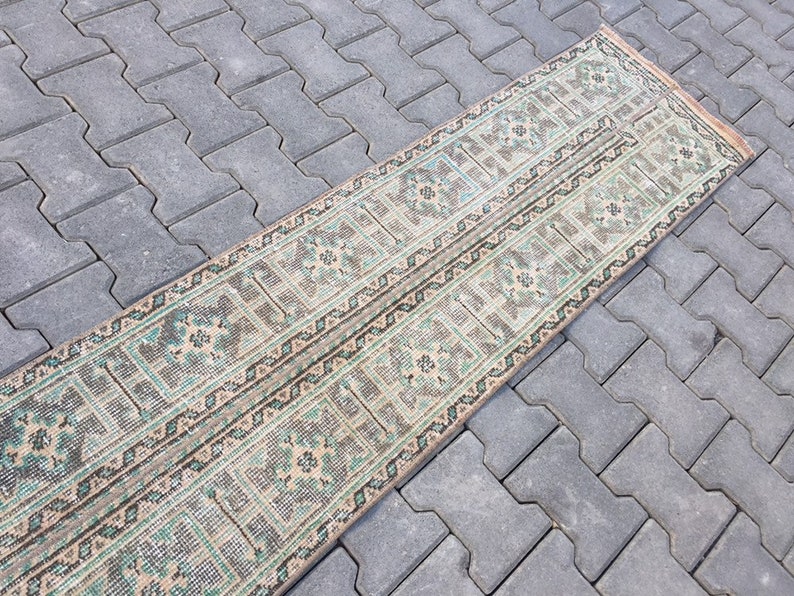 May include: A long, narrow rug with a geometric pattern in shades of beige, green, and brown. The rug has a distressed look, with some areas faded and worn.