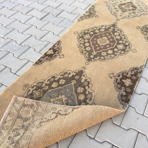 Pu&ograve; includere: Un tappeto da corridoio beige con un designo persiano tradizionale che presenta motivi intricati in tonalit&agrave; di marrone, beige e blu. Il tappeto &egrave; piegato a met&agrave;, rivelando il lato inferiore.
