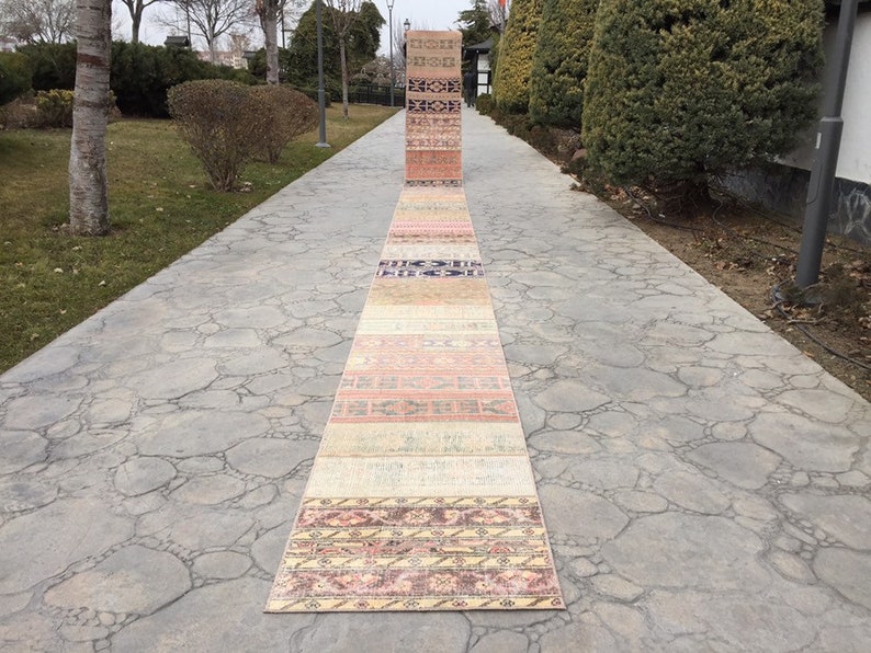 May include: A collection of colorful vintage rugs laid out on a stone walkway. The rugs are in various shades of pink, orange, yellow, and blue, with intricate patterns and designs.
