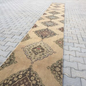 Pu&ograve; includere: Un tappeto lungo beige con un designo persiano tradizionale. Il tappeto presenta un medaglione centrale con intricati motivi floreali e un bordo di motivi ripetuti. Il tappeto &egrave; steso su una superficie di cemento grigia.