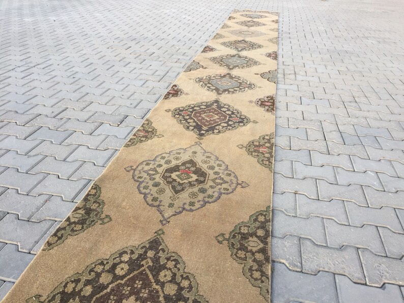 Pu&ograve; includere: Un tappeto lungo beige con un designo persiano tradizionale. Il tappeto presenta un medaglione centrale con intricati motivi floreali e un bordo di motivi ripetuti. Il tappeto &egrave; steso su una superficie di cemento grigia.