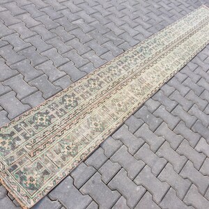 May include: A long, narrow rug with a traditional Persian design in shades of green, brown, and beige. The rug is laid out on a gray concrete patio.