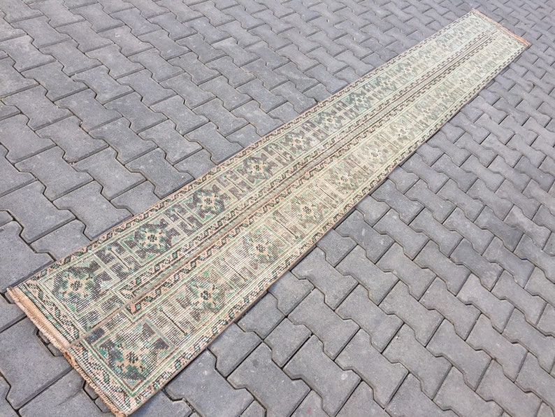 May include: A long, narrow rug with a traditional Persian design in shades of green, brown, and beige. The rug is laid out on a gray concrete patio.