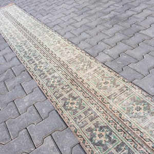 May include: A long, narrow rug with a traditional pattern in shades of beige, green, and brown. The rug is laid out on a gray concrete surface.
