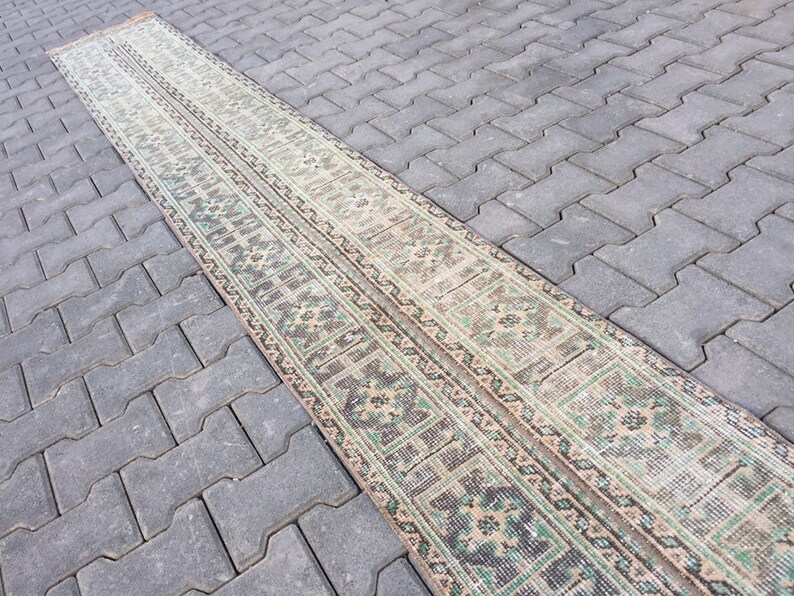 May include: A long, narrow rug with a traditional pattern in shades of beige, green, and brown. The rug is laid out on a gray concrete surface.