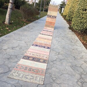 May include: A long, narrow, multi-colored rug with a geometric pattern. The rug is laid out on a gray concrete walkway.
