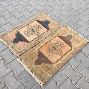 May include: Two small, rectangular rugs with intricate geometric patterns in shades of brown, beige, and green. The rugs have a fringed edge and are laid out on a grey concrete surface.