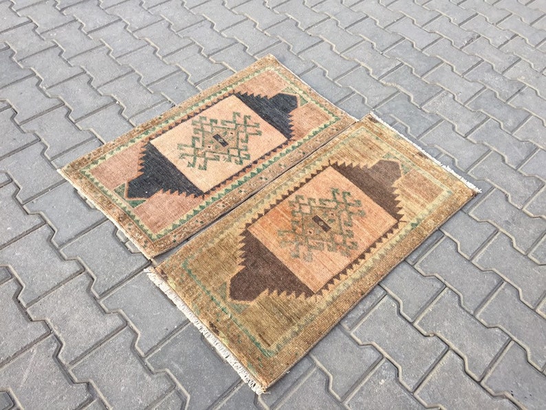May include: Two small, rectangular rugs with intricate geometric patterns in shades of brown, beige, and green. The rugs have a fringed edge and are laid out on a grey concrete surface.