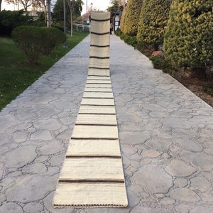 Puede incluir: Una alfombra larga y estrecha de color blanco roto con rayas marrones colocada en un camino de piedra. La alfombra está doblada por la mitad y tiene flecos en los extremos.