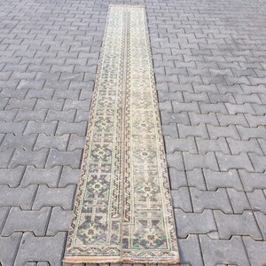 May include: A long, narrow rug with a cream background and a repeating pattern of green, brown, and black floral motifs. The rug is laid out on a gray concrete surface.