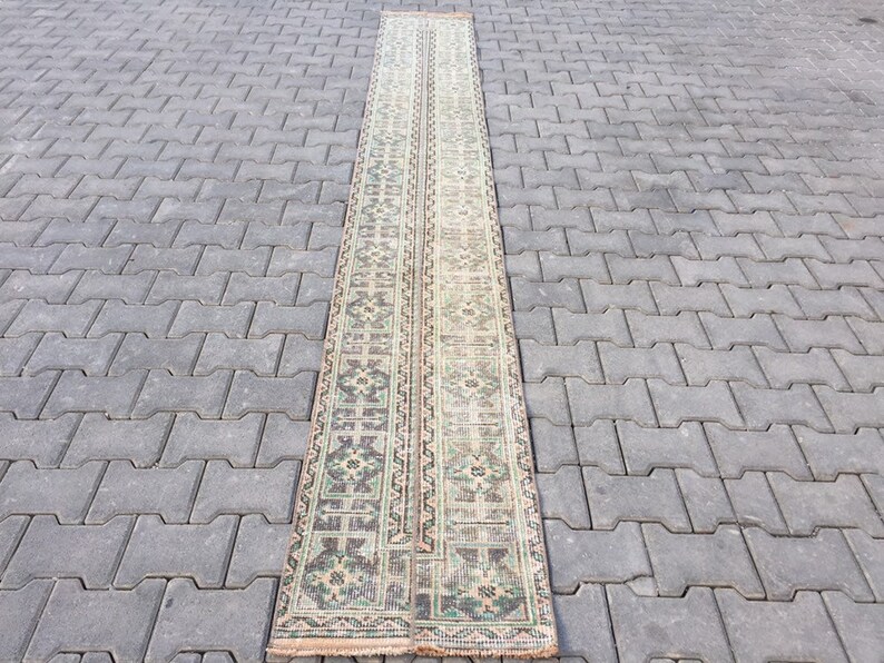 May include: A long, narrow rug with a cream background and a repeating pattern of green, brown, and black floral motifs. The rug is laid out on a gray concrete surface.