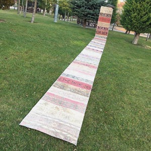May include: A long, narrow rug with a faded, vintage look. It has a repeating pattern of horizontal stripes in red, pink, beige, and brown. The rug is displayed outdoors on a green lawn, with trees in the background.