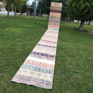 May include: A long, narrow rug with a colorful, geometric pattern. The rug is made of woven fabric and has a fringed edge. It is laid out on a grassy lawn.