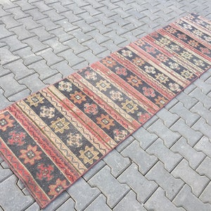 Op de afbeelding: Een lang, smal tapijt met een traditioneel geometrisch patroon in tinten rood, oranje, beige en zwart. Het tapijt is gemaakt van wol en heeft een verweerde, vintage uitstraling.