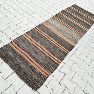 Puede incluir: Una alfombra rectangular tejida a mano, con una base marrón oscuro y rayas naranjas. La alfombra está hecha de fibras naturales y tiene un aspecto texturizado. La alfombra está colocada sobre una superficie de baldosas gris claro.